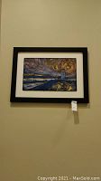 Framed landscape artwork featuring a lighthouse under a swirling night sky, displayed on a wall.