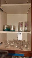 Overview of three art glass tumblers, two Havana Club glasses, and eight clear crystal shot glasses on cabinet shelf
