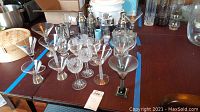 Wide view of assorted clear glass stemware, including martini glasses, cocktail glasses, and metal shakers in background.