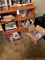 Wood bookshelf filled with assorted books, some on the floor and in a box adjacent to it.