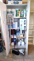 Cabinet filled with cleaning supplies including paper towels, mop, broom, various cleaning sprays and powders.