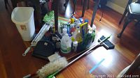 Photo showing assorted cleaning supplies on the floor including bottles, a brush, a feather duster, and a plastic waste bin.