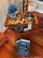 Photo showing an assortment of decorative items on a wooden tray atop a side table with wooden legs. Items include a large decorative plate with a sailing ship design, two ceramic bowls each holding black crustacean figures, stones, geode bookends, and a tall glass vase with decorative sticks. Additional items are displayed on the lower shelf and floor below the table.