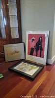 Three framed artworks leaning against a wall on hardwood floor. One smaller sepia-tone drawing, one vertical poster of horse and rider, and one horizontal colorful drawing.