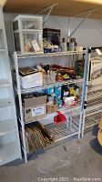 Wide view of a white metal wire shelving unit holding various hardware, tape, locks, lightbulbs, lubricant spray cans, leaf bags, and other items stored in boxes and small plastic drawers.