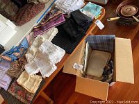 Photo showing various cloth napkins, placemats, and tablecloths spread out on a table with a box containing more linens.