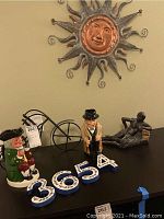 Photo of assorted decorative items on black table including Royal Doulton Churchill jug, house numbers, reclining nude sculpture, black metal bicycle bottle holder, and metal sun wall hanging on wall.