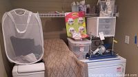 Wide view of storage area showing clothes basket, rug runner over white mini fridge, small drawer unit with Command wall hangers, plastic storage containers, and handheld vacuum.