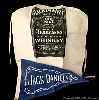 Off-white apron with large black Jack Daniel's Old No. 7 label printed on the front, paired with a blue promotional pennant with white script.