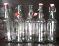Five clear glass bottles standing upright in a row, each with a metal swing lock cap featuring white and red tops, on a dark surface.