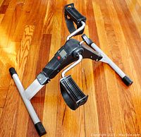 Side angle view of the TABEKE pedal exerciser showing the black pedals with straps, silver metal frame, and digital display unit.