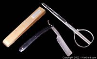 Vintage straight razor with black handle, metal blade, showing wear and patina, next to metal barber scissors and beige case on black background.