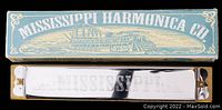 View of the vintage Mississippi Harmonica Co. metal harmonica with engraving, placed below its original rectangular cardboard box featuring a steamboat illustration and the company name in blue and white.