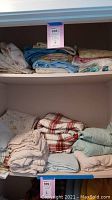 Shelf one with folded bed linens including light blue sheets, off-white sheets, plaid and floral patterns.