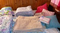Overview of folded blankets, pillows, and new packaged sheets on bed with wood paneling background
