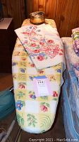 Full view of ironing board with floral and fruit patterned cover, tin container on top with buttons, and folded vintage floral tablecloth.