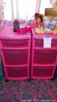 Two pink plastic storage units side by side, each with three drawers, with dolls and items on top