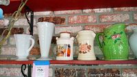 Five decorative vases and pitchers displayed on a shelf with a brick background, showing milk glass and ceramic pieces with floral patterns