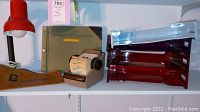 Photo showing arrangement of red metal desk lamp, vintage ledger binder, Rolodex, plastic file trays, and wooden shoehorn or letter opener.
