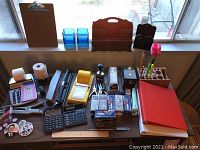 Wide shot of the entire collection of office supplies on a table in front of a window showing clipboards, calculators, staplers, binders, organizers, buttons, pens, and other stationery items.