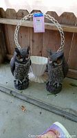 Two gray plastic owl statues and a white planter with lattice frame photographed outdoors on concrete against a wooden fence.