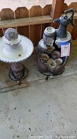 Photo showing two patio fountains side by side on a concrete floor by a wooden fence. Left fountain has a ball atop a scalloped dish. Right fountain has water pump handle on top with multiple basins on barrel base. Electric cords visible.