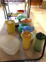 Several plastic food storage containers in various colors and sizes including pitchers and bowls, lined up on a floor.