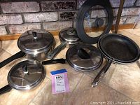 Photo showing collection of stainless steel pots with black handles, several stainless lids, a frying pan, and a glass lid, all on tiled surface.