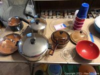 Multiple pots and pans with lids, glass and wooden bowls, plastic cups, and various kitchenware items arranged on a table.