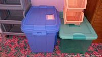 Two large plastic lidded storage bins, one blue and one green, positioned side by side on a multi-color carpet next to wooden furniture.