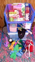 Two stacked blue plastic storage bins filled with children's read-along books, a miniature green metal bucket on top, knitted pink tissue box cover, black gloves, colorful knitted gloves, and a Hello Kitty cup in a clear red plastic case, plus various other small toys and items arranged in front.