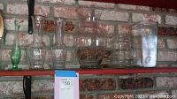 Wall shelf with seven glass vases of varied designs including clear and green glass. Shelf against a brick wall.