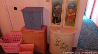 Photo showing four hampers—pink plastic laundry hamper, peach-colored hamper, large orange hamper, pink vintage metal hamper—and two framed child-themed prints against a pink wall and next to a cream-colored waste basket.