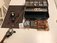 Full lot display photo showing the green metal tackle box open with assorted fishing lures and accessories inside, fishing rod, and reel.