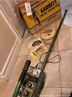 Full view showing complete Garrett metal detector in green color with attached coil, manual, and original box on tile floor.