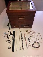 Wooden jewelry box with glass top and 4 drawers, assorted fashion jewelry and 2 wristwatches with black straps laid out in front