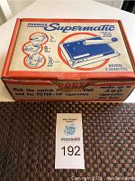Top view of original box of Premier Supermatic cigarette making machine showing name, machine illustration, instructions, and claims to make up to 300 cigarettes per hour.