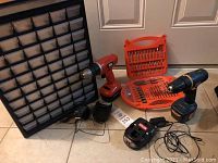 Two cordless drills, battery chargers, orange plastic drill bit cases, and black plastic organizer with drawers, all items shown together on floor.