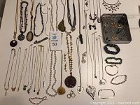 Photo showing a collection of various necklaces, bracelets, and earring sets arranged on a flat surface including metal chain styles, beaded jewelry, and pendant necklaces.