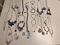 Fashion jewelry displayed on white surface including necklaces, bracelets, and earrings varied in color and style. Several necklaces with pendants, matching earrings, and bracelets laid out.