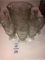 Full view of 9 glasses arranged around one matching pitcher, clear glass shows floral reliefs and fluted bases.