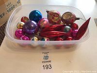 Top view showing an assortment of vintage glass Christmas ornaments in a plastic container including round and elongated shapes in pink, blue, purple, red, and gold hues with painted and glitter accents.