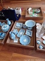 Overview of seven Wedgwood Jasperware pieces including vases, dishes, lidded boxes, and a pedestal bowl in matte blue with white relief detailing, arranged in cardboard trays.