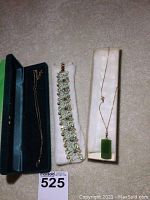 Photo showing three jewelry pieces: a gold chain necklace with green pendant, an ornate bracelet with stones, and a gold chain necklace in a box.