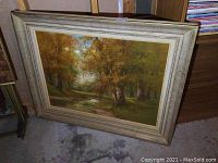 Overall view of oil painting in textured off-white frame, showing landscape with trees and stream.