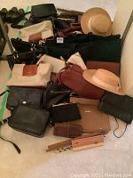 Full view of all items in the lot showing multiple handbags, purses, wallets, two straw hats, and a wooden tie hanger on carpeted floor.