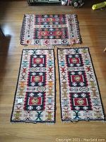 Photo showing all three rugs laid out on the floor; one larger rug at top with multicolor rectangular motifs and fringe, and two longer narrow rugs below with repeating square and geometric designs and fringed edges.