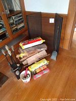 Photo showing black metal fireplace screen, wood stand with four brass-handled fireplace tools (poker, brush, shovel, tongs), two packages of Duraflame logs, assorted wood pieces, and bucket with accessories and matches.