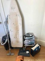 Full lot view showing ironing board, iron on box, Kenmore stick vacuum with hose, wood stool and Eureka hand vacuum base and accessories