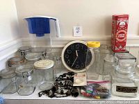 Photo of vintage glass jars, Brita water filter pitcher, round black wall clock, red metal tea tin, black metal trivet, windchime parts, and small miscellaneous items.